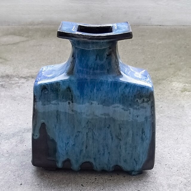 A square, blue ceramic vase with a narrow neck and flat rim, featuring a glossy, drip glaze finish, sits on a concrete surface.